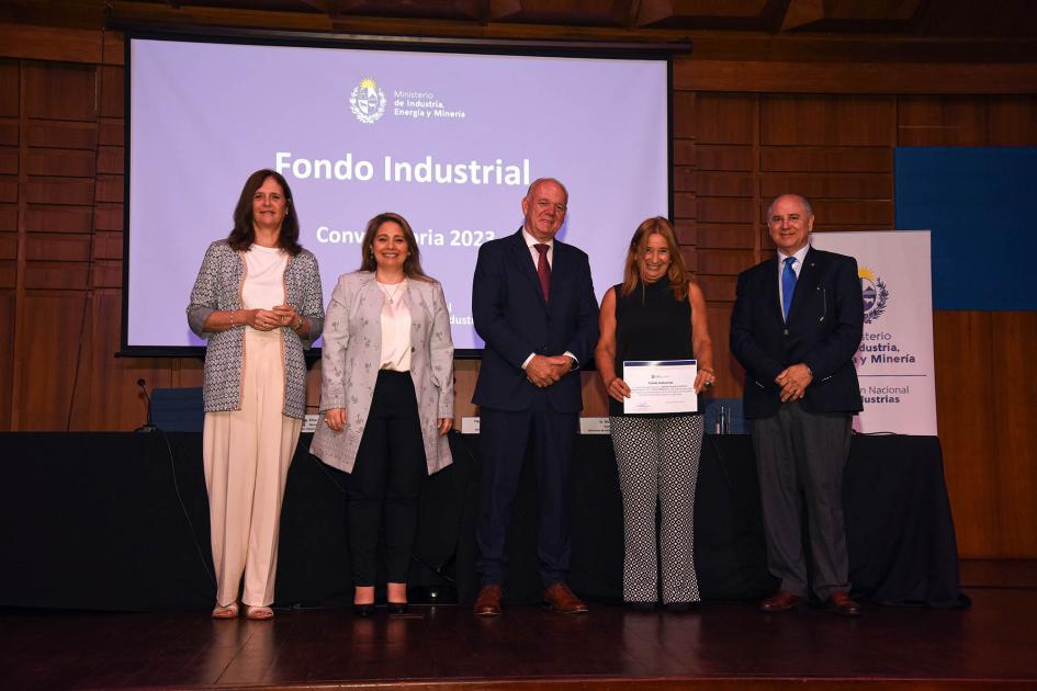 Autoridades acompañan a una mujer que muestra su diploma; detrás hay una pantalla