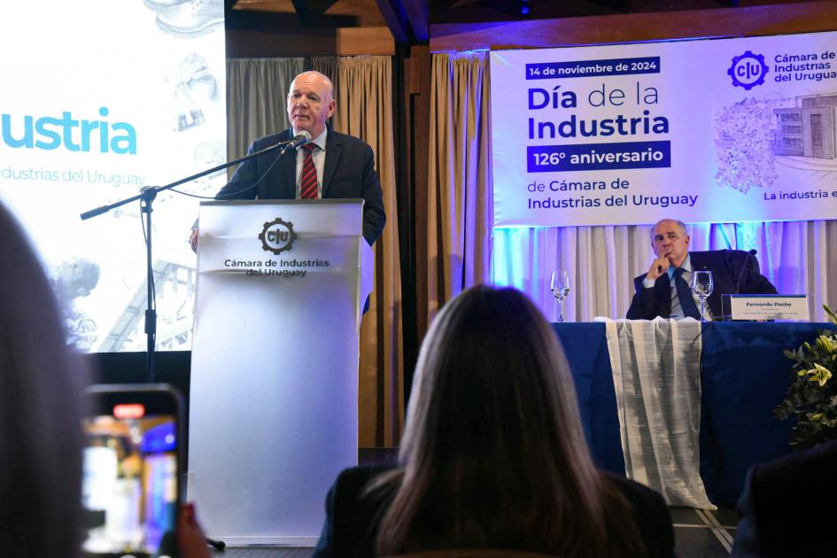 El ministro interino Walter Verri expuso en el Día de la Industria Verri habla dese un atril ante el público; al lado, Pache, sentado; detrás, pantalla