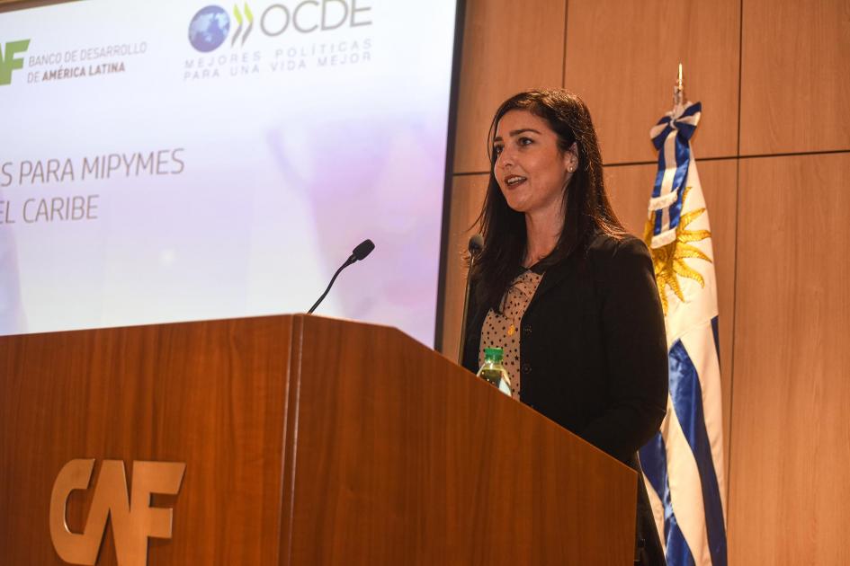 Coordinadora de Relaciones Internacionales del MIEM, Celeste Elhordoy Coordinadora de RR. II. del MIEM actúa como maestra de ceremonias en un atril; detrás, bandera nal.