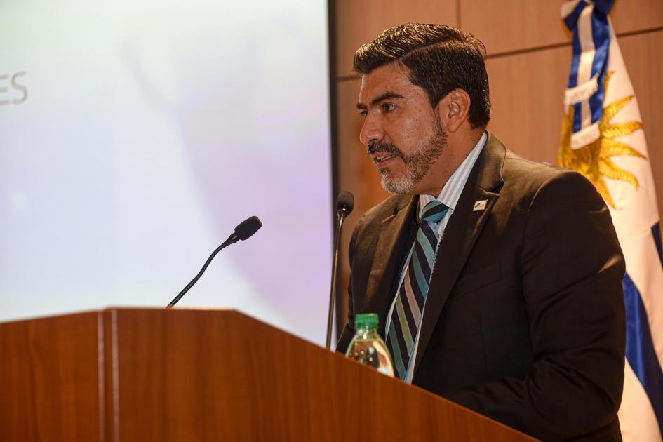 Emb. Clarems Endara, secretario permanente del Sistema Económico Latinoamericano y del Caribe (SELA) En un atril habla Clarems Endara, secretario permanente de SELA; detrás, la bandera uruguaya