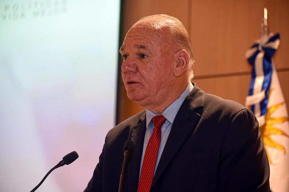Ministro interino Walter Verri Habla Walter Verri con un micrófono; detrás, la bandera uruguaya y un fragmento de pantalla