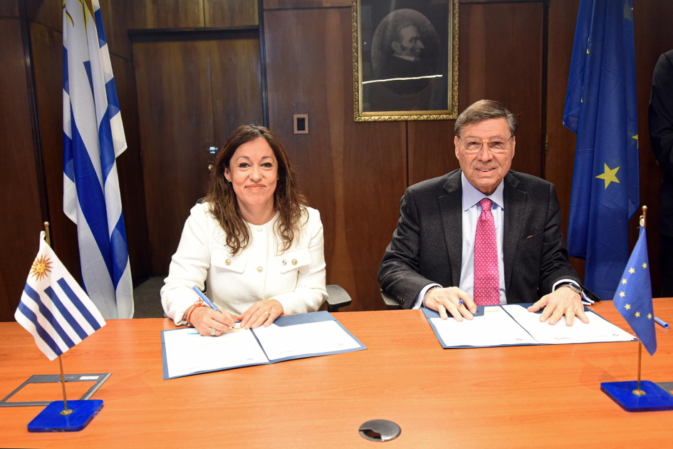 Ministra Fernanda Cardona y presidente de RedCLARA, José Manuel Palacios Cardona y Palacios, sentados, con lapicera y documento; detrás, bandera uruguaya