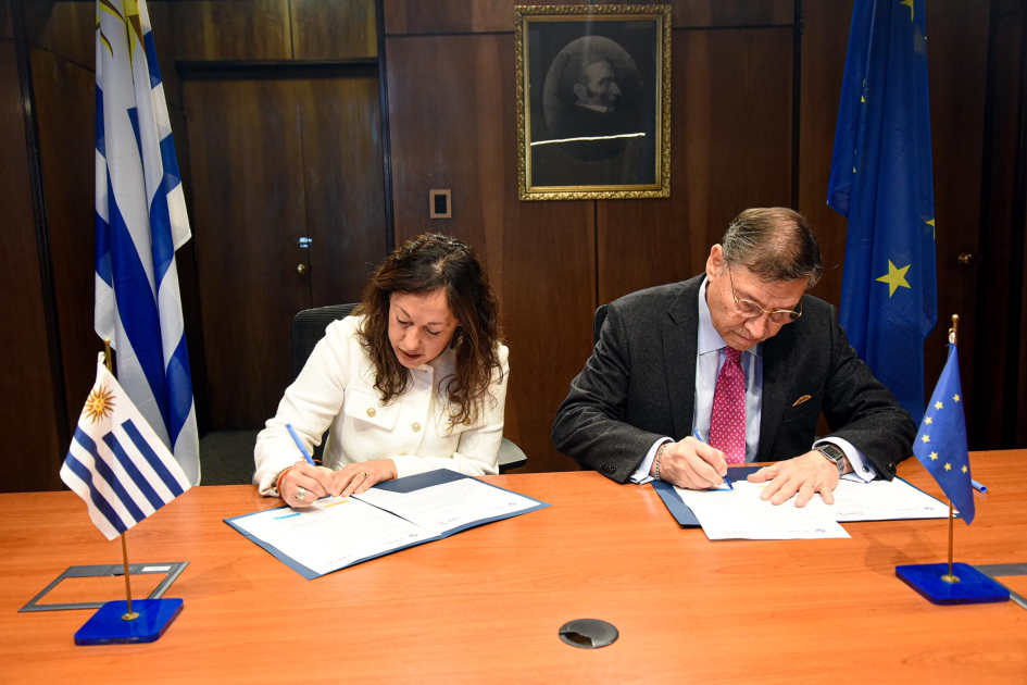 Ministra Fernanda Cardona y presidente de RedCLARA, José Manuel Palacios Cardona y Palacios firman documentos en una mesa; detrás, bandera uruguaya