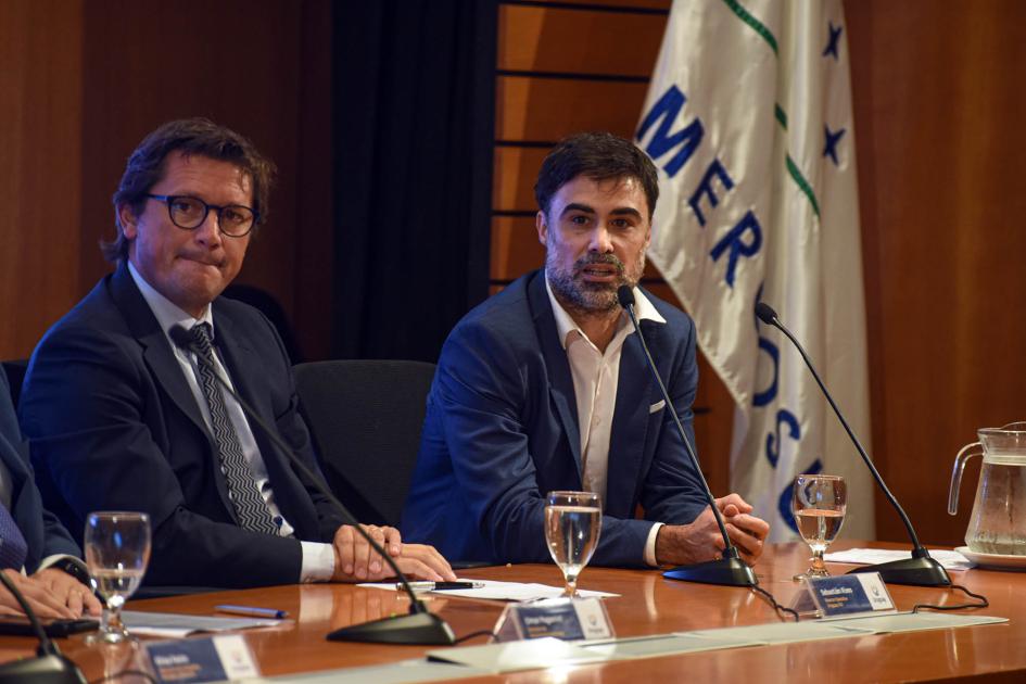 Director de Laboratorio Metodológico de Equipos Consultores, Santiago Peyrou Peyrou habla junto a Risso; a su lado, una bandera del Mercosur
