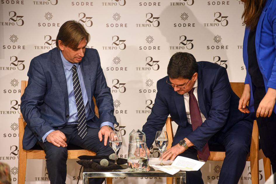 Firma del convenio Cantero firma en una mesa baja; junto a él está Delgado; a su izquierda, se ve una mujer (fragmento)