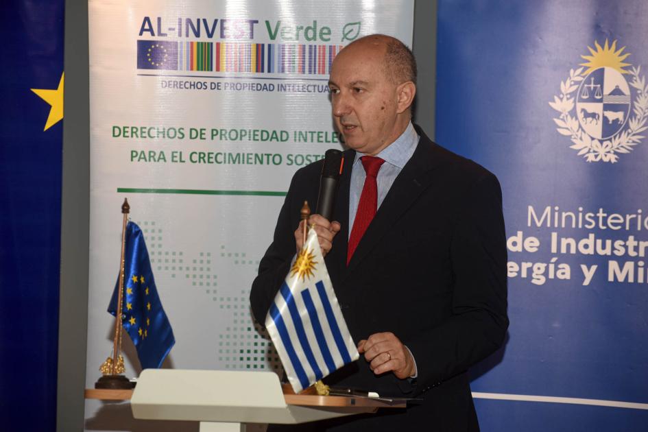 Berizzi habla; en un atril, banderas uruguaya y de la Unión Europea; detrás, banners