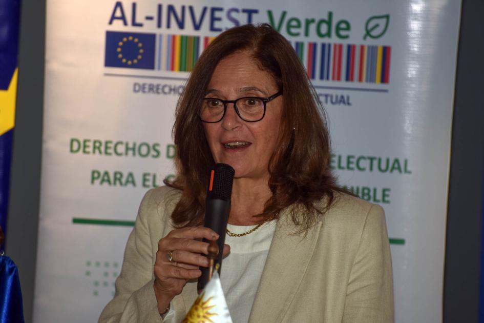 Ministra Facio habla con micrófono; detrás, banner de AL-INVEST Verde