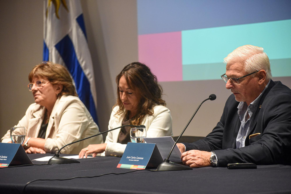 Mesa de autoridades Canessa habla junto a Cardona y Fernández; detrás, pantalla y bandera uruguaya