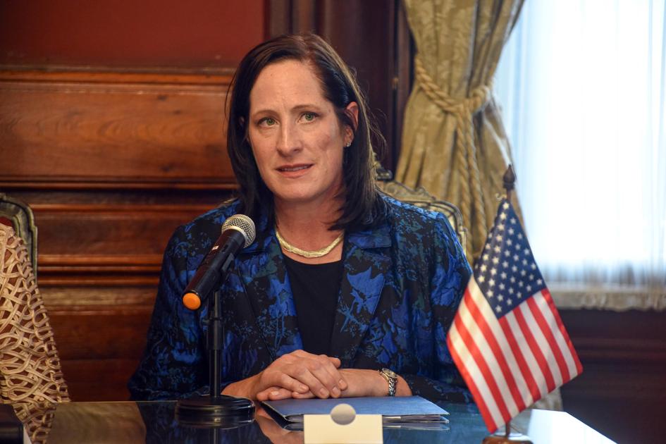 Embajadora de Estados Unidos habla; frente a ella, en la mesa, una bandera de Estados Unidos