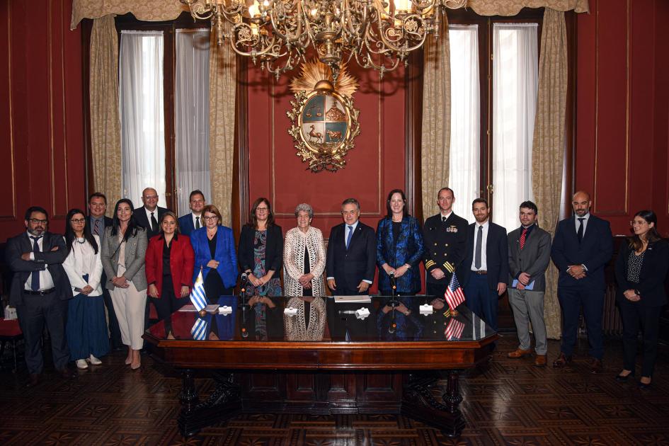 Autoridades y funcionarios posan de pie; frente a ellos, una mesa; detrás, antiguo escudo uruguayo