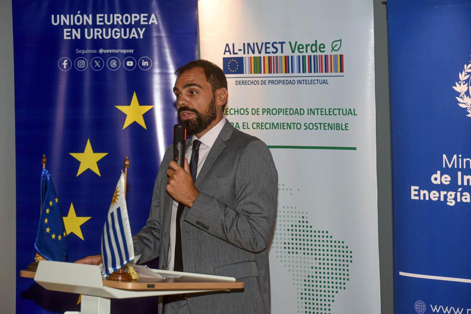 Director Martínez habla con un micrófono desde atril con banderas uruguaya y de UE; detrás, banners
