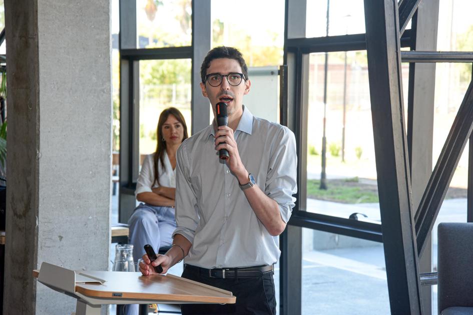 Federico Wyss, consultor en Competitividad, Tecnología e Innovación del BID Wyss habla con un micrófono; detrás, maestra de ceremonias