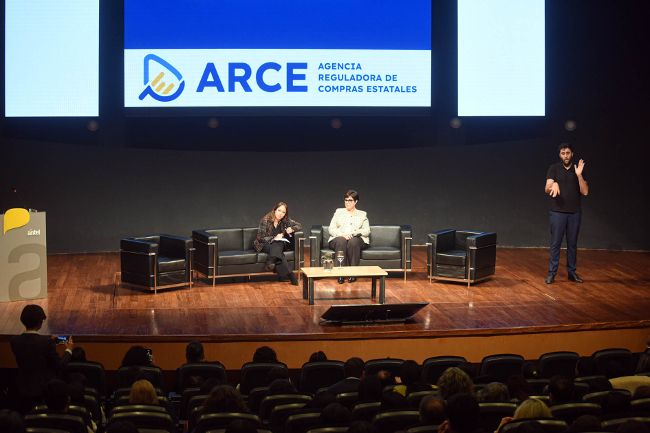 Cierre de la VIII edición de las Jornadas Técnicas de Compras y Contrataciones Estatales Cardona y Burguez, sentadas en un escenario; más cerca, público; detrás, pantalla con logo de ARCE