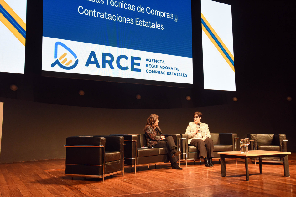 Ministra Fernanda Cardona y directora de ARCE, Isis Burguez Cardona y Burguez, sentadas en un escenario; más cerca, público; detrás, pantalla con logo de ARCE