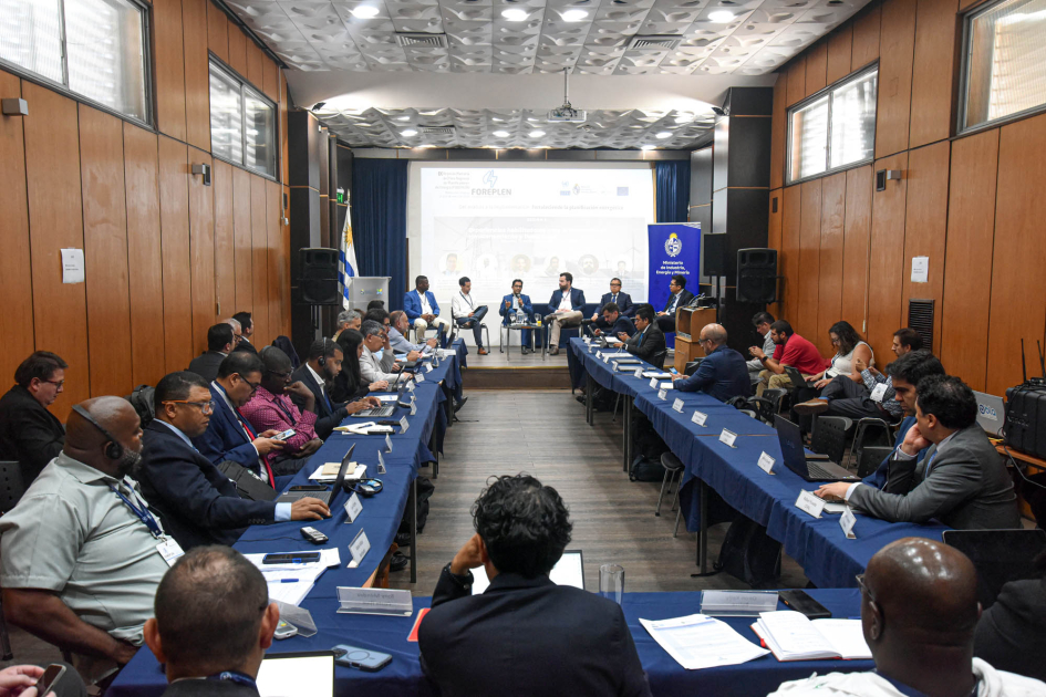 Personas sentadas en mesas en forma de "u"; al fondo, personas exponen en un escenario