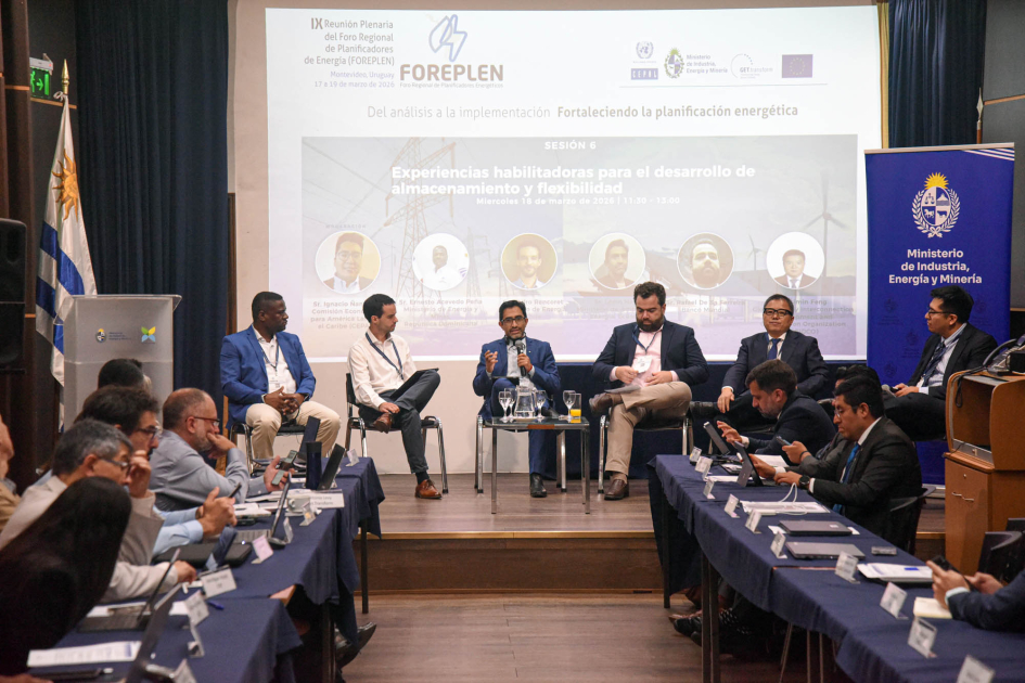 Personas sentadas en un escenario; detrás, pantalla; frente a ellas, personas en mesas enfrentadas