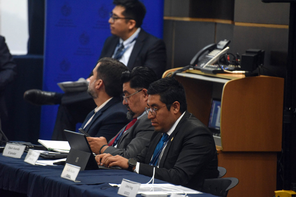 Personas sentadas en una mesa; detrás, hombre sentado en segunda fila