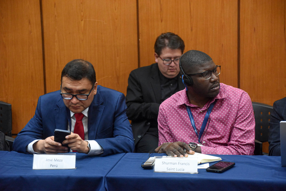 Dos hombres, sentados en una mesa; detrás, hombre en segunda fila de participantes