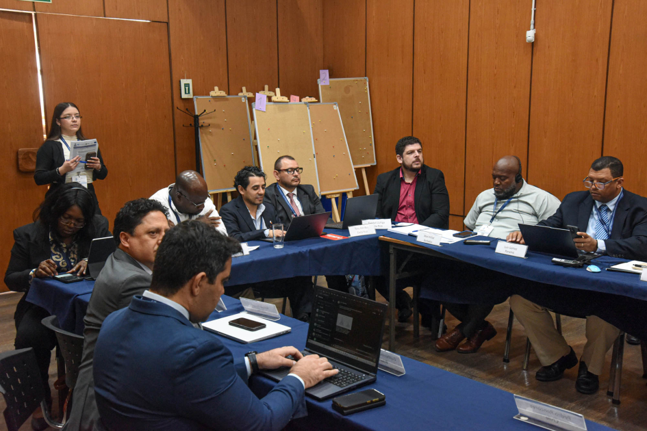 Personas sentadas en mesas en forma de "u"; detrás, mujer de pie