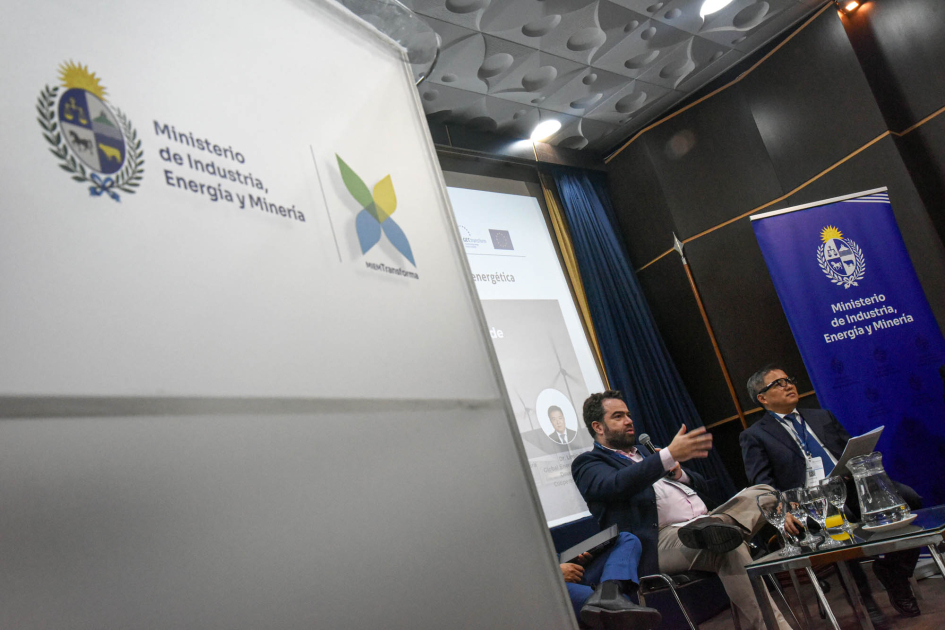 Al fondo, personas en un escenario y banner del MIEM; más cerca, atril con logos del MIEM