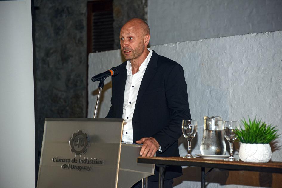 Funcionario del MIEM y presentador del evento, Venancio Lacurcia Maestro de ceremonias habla desde un atril; a su lado, una mesa con copas y una planta