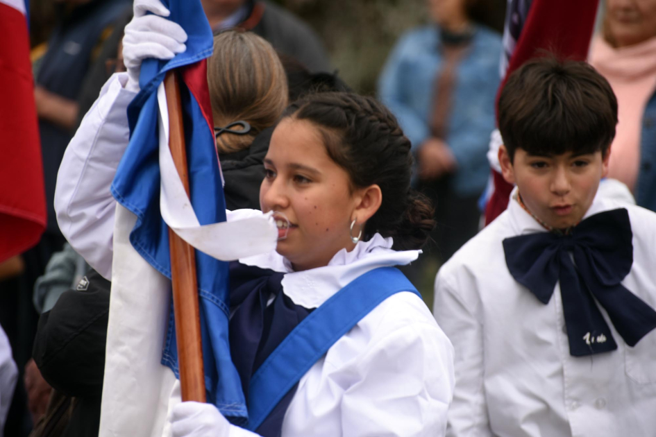Niña de túnica lleva bandera. Detrás niño de túnica y moña