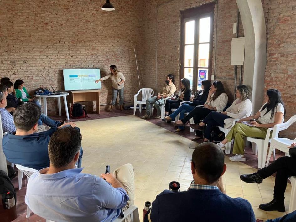 Una mujer presenta datos en una pantalla a un grupo de personas sentadas en ronda en un salón