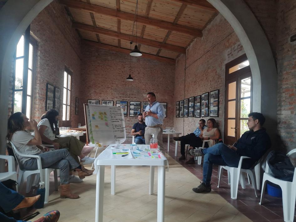 Un hombre de pie, en el centro de un salón, dialoga con personas sentadas; hay una mesa