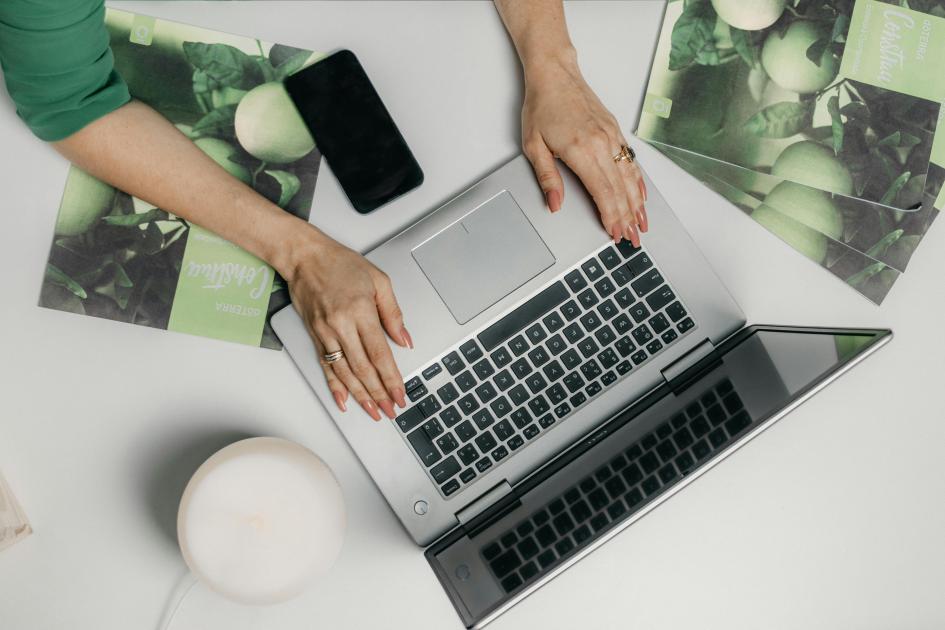 Manos de mujer escriben en una laptop; en la mesa hay fotos que parecen diseños (vista desde arriba)