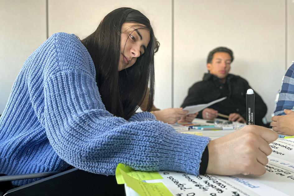 Una adolescente o joven escribe en una hoja; a lo lejos, se ve otro