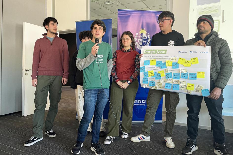 Adolescentes y jóvenes, de pie; algunos sostienen un gran cartel; otro, un micrófono