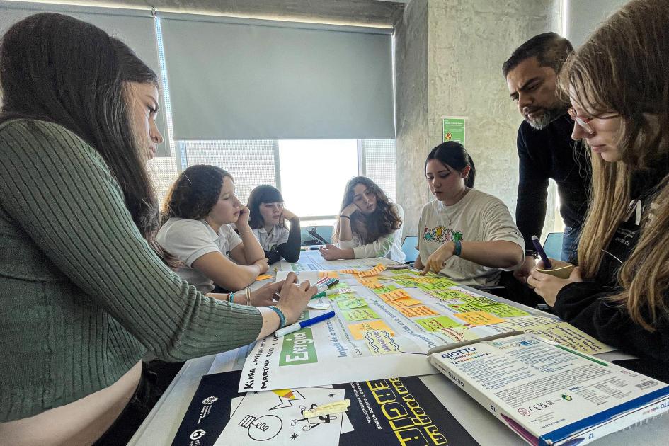 Jóvenes trabajan con papeles y lapiceras en una mesa; un adulto está de pie