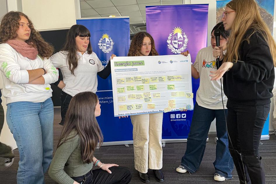 De pie, un grupo de adolescentes sostiene una gran cartulina; detrás, banners institucionales
