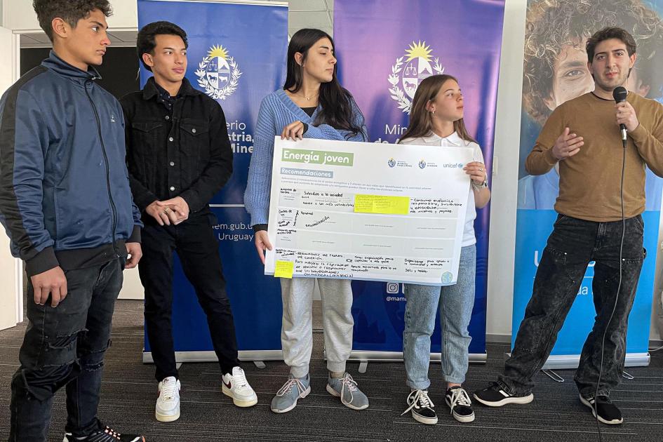 Adolescentes y jóvenes; dos sostienen un cartel; otra persona, un micrófono