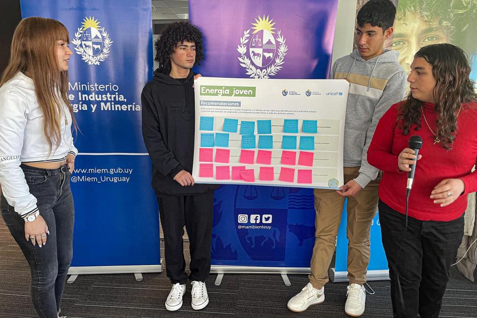 Adolescentes participantes; dos sostienen un gran cartel; otra, un micrófono; detrás, banners