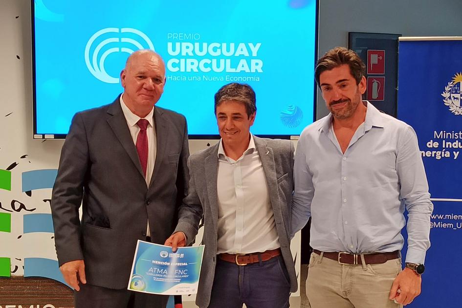 El subsecretario Walter Verri entrega una mención Verri entrega una mención a un hombre que la sostiene en su mano; a su lado hay otro hombre