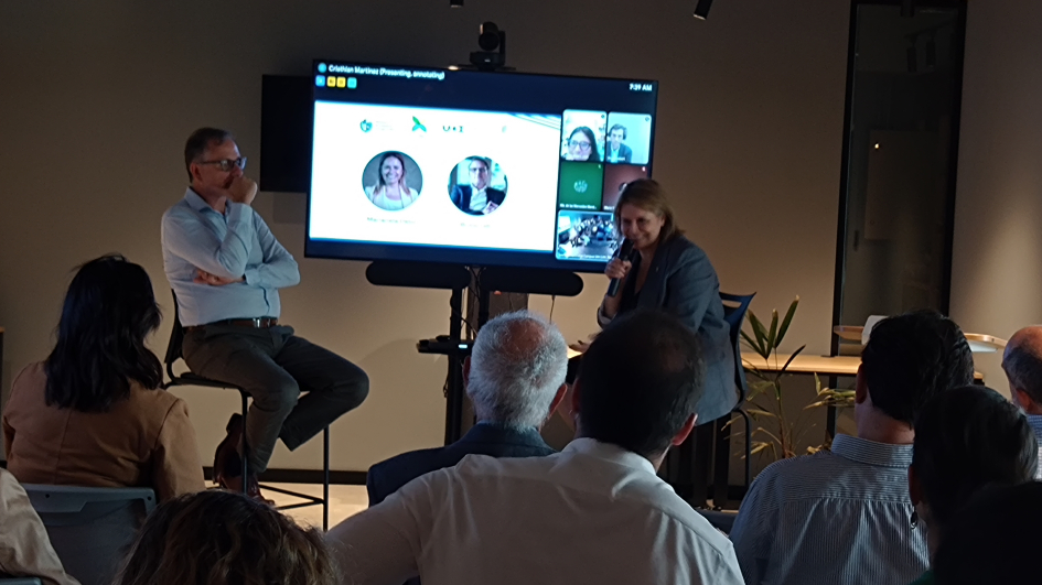 Presentación Bruno Gili y Marianela Delor 2 personas sentadas frente a un tv y gente mirándolos