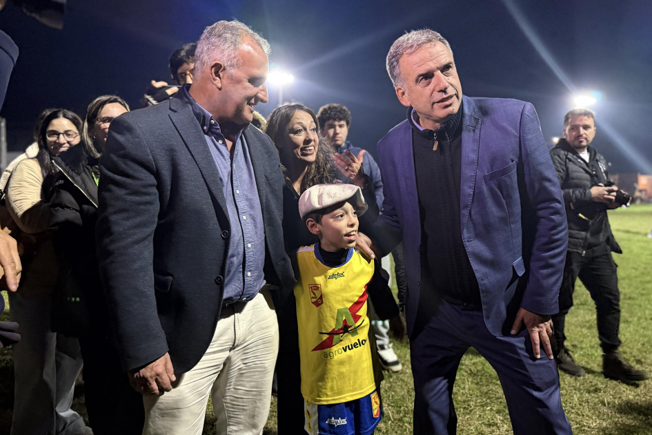 Inauguración de luminarias en la cancha de baby fútbol de José Pedro Varela Orsi, Cardona y Ximénez, junto a un niño con boina, en una cancha iluminada