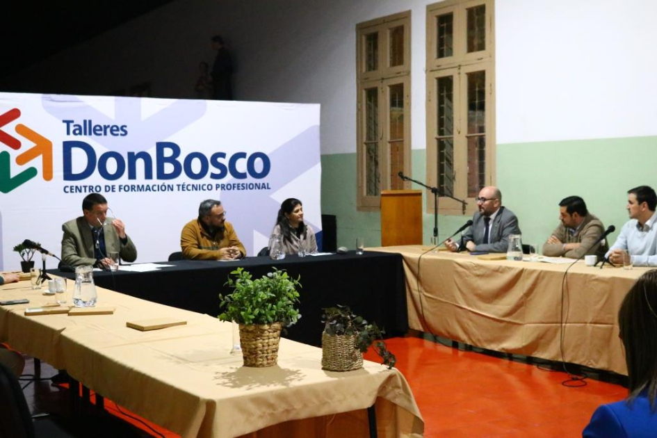 Personas sentadas en mesas en forma de "u"; al fondo, banner de Don Bosco