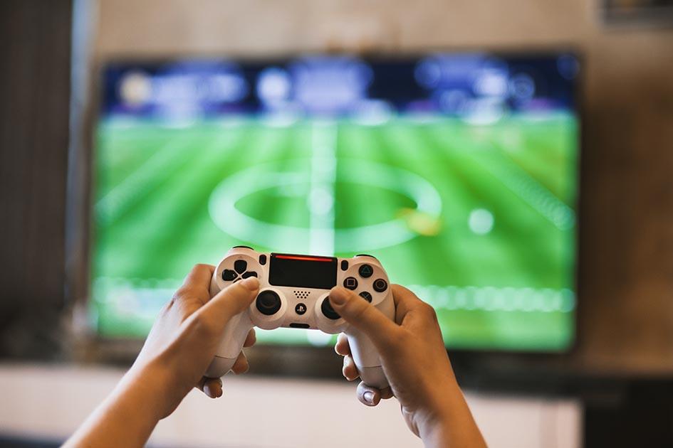 Foto de manos que juegan con un joystick frente a un televisor con una cancha de fútbol