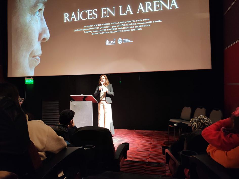 Una mujer habla en un escenario; encima, una pantalla