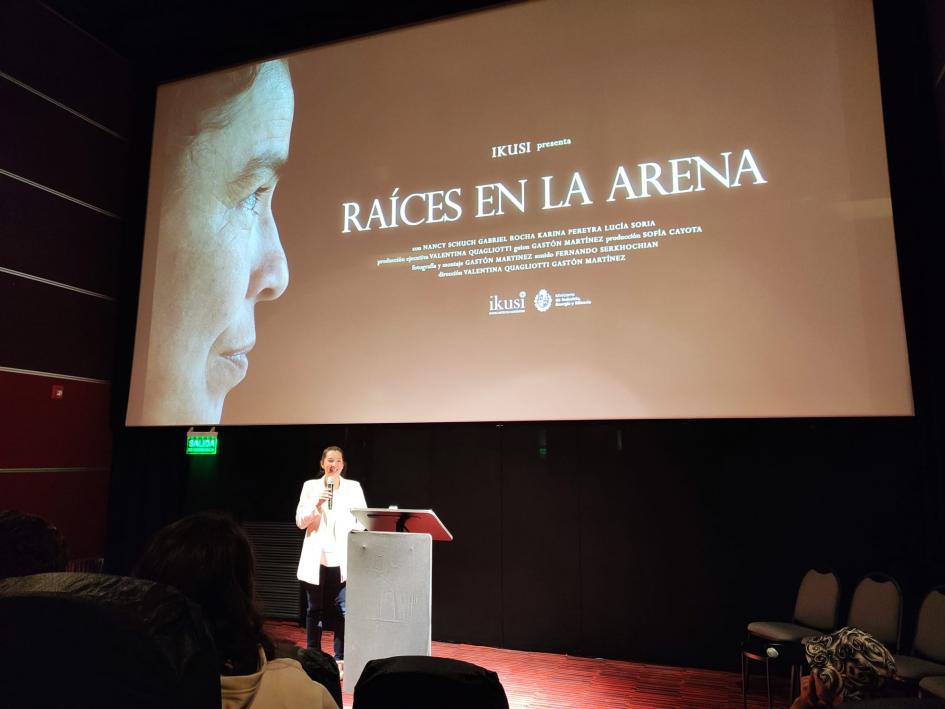 Palabras de la directora de IKUSI, Valentina Quagliotti Una mujer habla desde un escenario; encima, una gran pantalla con el corto