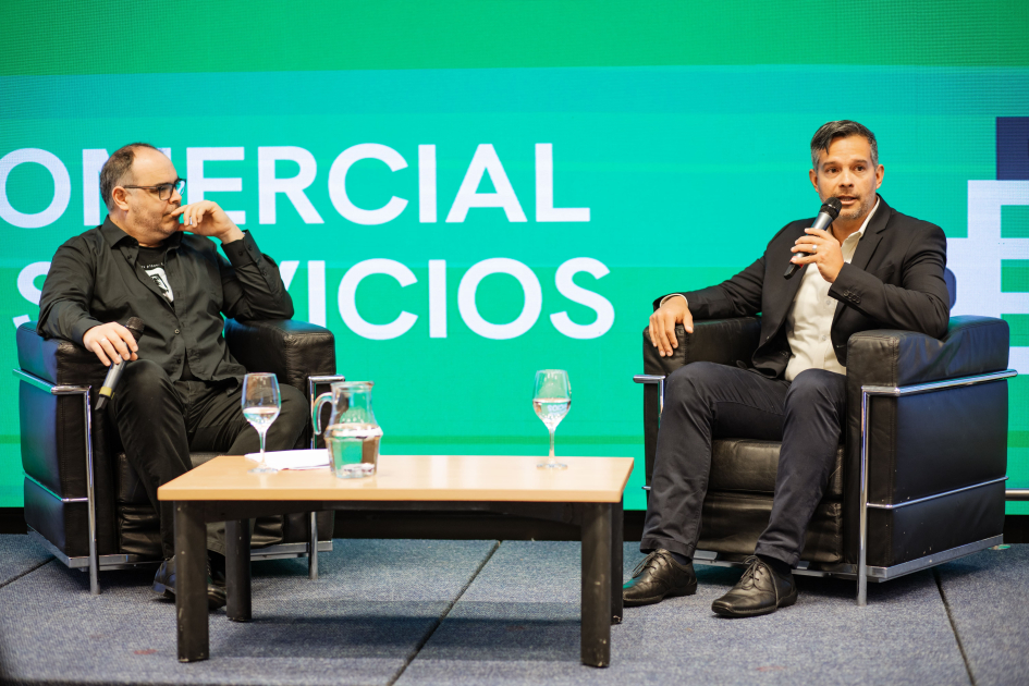 Dos hombres hablan en dos sillones; detrás, pantalla