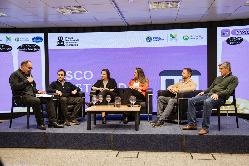 Los cinco representantes de ESCO, sentados junto a presentador, intercambian; detrás la pantalla
