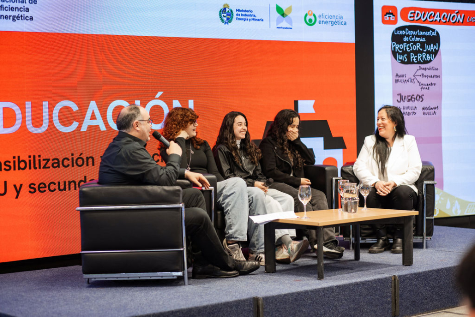 Estudiantes, docente y presentador, sentados en living, intercambian; pantalla detrás