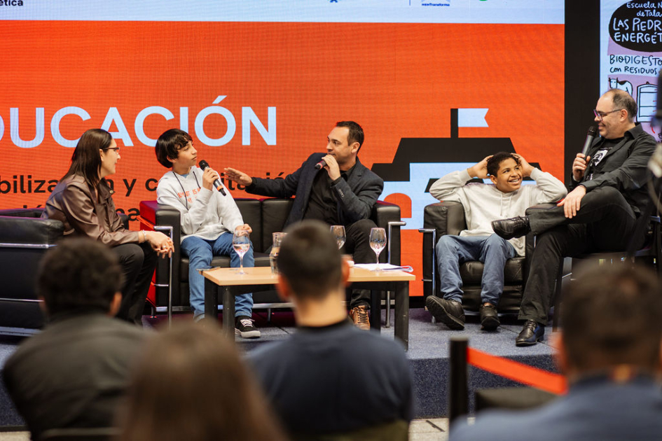 Estudiantes, docente y presentador, sentados en escenario, intercambian; detrás pantalla