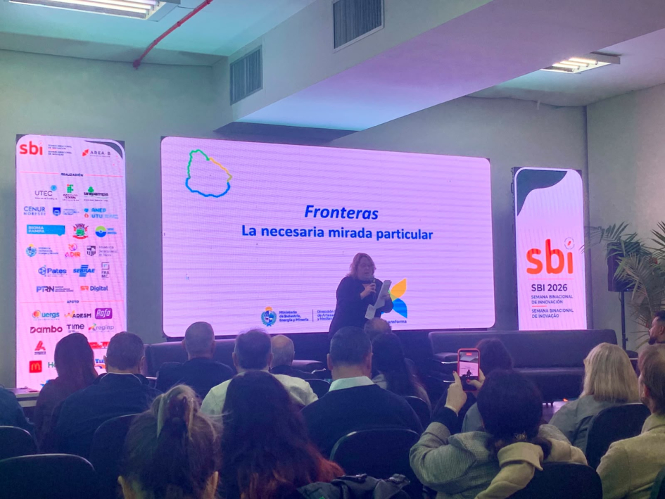 Directora Cidrás, de pie; detrás, cartelería; más cerca, público Ximena Cydras exponiendo en la semana binacional de la innovación