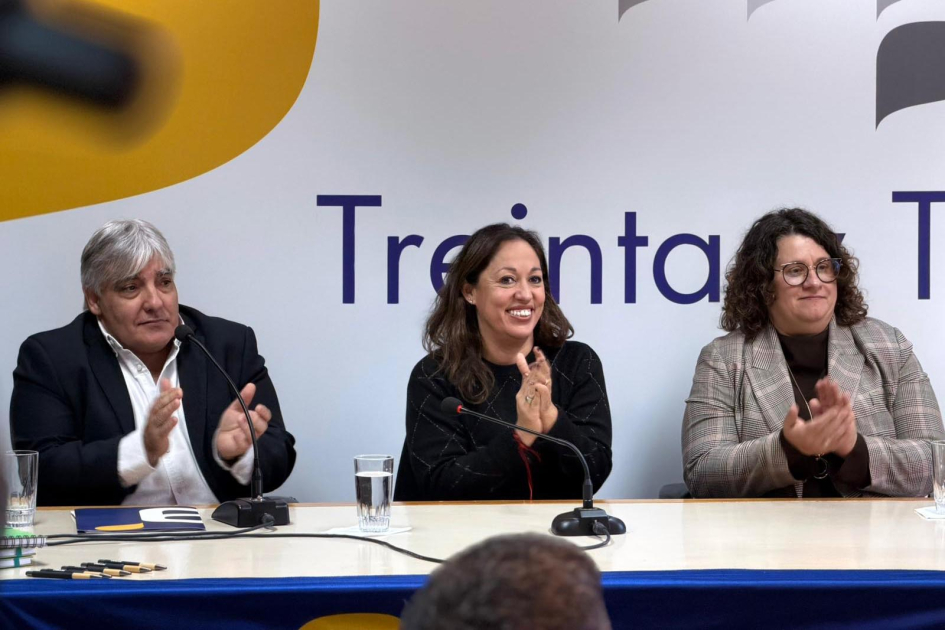 Ministra Fernanda Cardona, junto a Alejandro Paz (Antel) y Beatriz Martínez (Accesa) Cardona, Paz y Martínez aplauden, sentados; detrás, banner