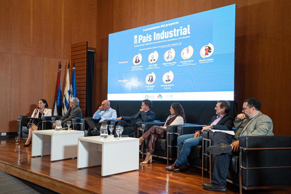 Expositores y expositoras, sentados en un escenario; detrás, pantalla