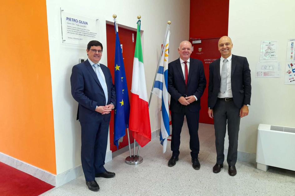 Reunión en el Campus Universitario de Savona Verri, Crego y otro hombre están, de pie, junto a las banderas de Uruguay, Italia y la UE